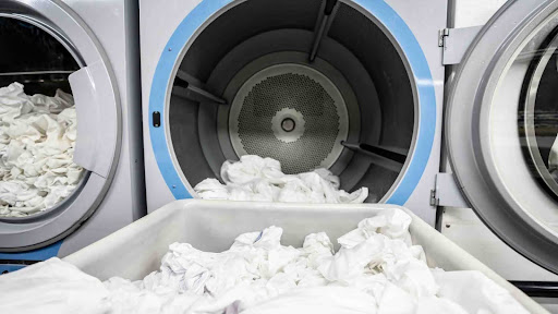 Shows commercial washing machines with one open and in process of filling with linens.